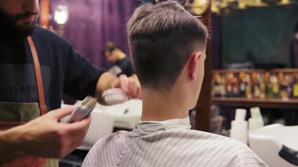 Vista posteriore di un cliente che si fa tagliare i capelli da un barbiere. Il parrucchiere barbuto taglia i capelli usando il trimmer elettrico — Video Stock