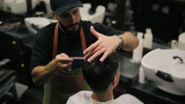 Barbeiro barbudo em avental pentear o cabelo de um cliente masculino sentado na cadeira na barbearia durante o corte de cabelo. Cabeleireiro elegante trabalhando em uma barbearia retro — Vídeo de Stock