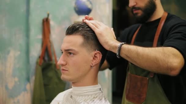 Вид сбоку на молодого красивого кавказца с пирсингом в ухе, одевающего волосы и стилизованного бородатым парикмахером в ретро-стильной парикмахерской. Слоумоушн — стоковое видео