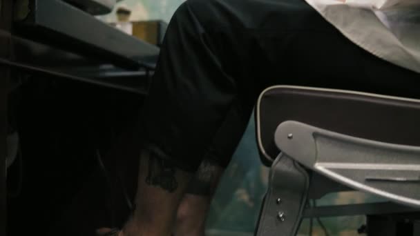 Jovem atraente com piercing e tatoo sentado em uma cadeira enquanto barbeiro barbudo está preparando-o para um corte de cabelo. Barbearia elegante. Movimento da câmera de suas pernas para cima — Vídeo de Stock