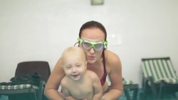 Χαριτωμένο ξανθιά νήπιο είναι καταδύσεις κάτω από το νερό μαζί με τη μητέρα του σε ειδικά προστατευτικά γυαλιά στην πισίνα. Η μητέρα του τον διδάσκει πώς να κολυμπήσετε. Ένα υποβρύχιο πυροβολισμό — Αρχείο Βίντεο