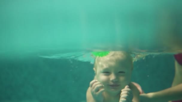 Criança loira bonito está mergulhando sob a água na piscina para obter o seu brinquedo, enquanto sua mãe está ensinando-o a nadar. Um tiro subaquático — Vídeo de Stock