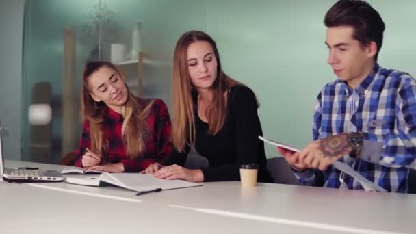 Tres estudiantes trabajando en sus tareas sentados juntos en la mesa y tomando café. Grupo de jóvenes en el encuentro en apartamento moderno. Disparo en cámara lenta — Vídeos de Stock