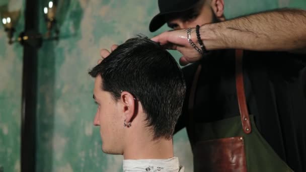 Un estilista peina suavemente el cabello corto y húmedo del cliente masculino. Peluquero barbudo en delantal peinando el cabello de un cliente masculino sentado en la silla en la barbería — Vídeos de Stock