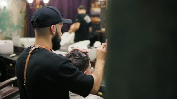 Vue latérale et arrière du coiffeur élégant avec barbe effectuant une coupe de cheveux avec des ciseaux tenant une brosse à cheveux dans ses mains. Mouvement lent de la caméra — Video