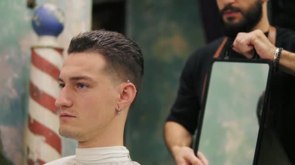 Cabeleireiro profissional mostra a visão traseira do corte de cabelo usando espelho para bonito cliente masculino satisfeito em uma barbearia de cabeleireiro profissional — Vídeo de Stock