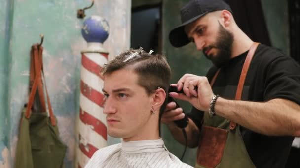 Joven hipster serio cortado por peluquero profesional en la peluquería. Peluquero barbudo joven de pie y haciendo corte de pelo elegante de hombre atractivo con clipper en la barbería — Vídeos de Stock