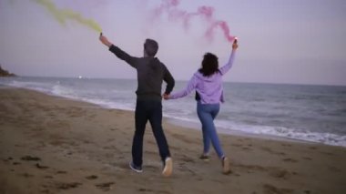 Arkadan görünüşü ellerinde renkli sis bombası ile deniz kenarında günbatımı sırasında çalışan genç bir çift. Çevrelerindeki eğlenceli, renkli duman
