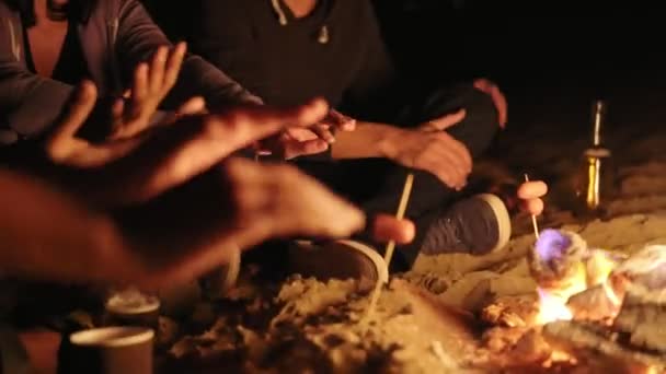 Picnic de jóvenes sentados junto a la hoguera en la playa y calentando sus manos por la noche. Vista de cerca — Vídeos de Stock