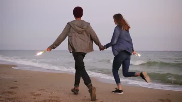 Vista posterior de la joven pareja multiétnica corriendo por el mar y sosteniendo velas brillantes durante el atardecer. Disparo en cámara lenta — Vídeos de Stock