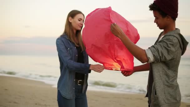 年轻的多民族夫妇举行红纸灯笼发射前。海滩上的浪漫约会漂亮的女人和她的非洲男友一起拿着火灯笼, 让它飞 — 图库视频影像