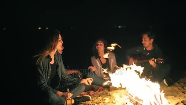 Jovens pessoas alegres cantando canções na fogueira tarde da noite, tocando guitarra, bebendo cerveja. Amigos alegres conversando e se divertindo — Vídeo de Stock