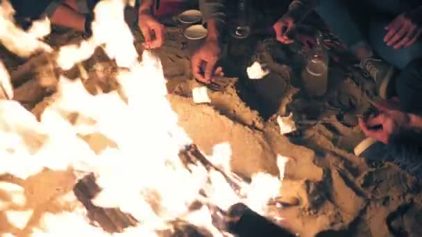 Vue du haut : mains tenant des bâtonnets avec des guimauves et les faisant frire la nuit. Groupe de personnes assises près du feu tard dans la nuit 