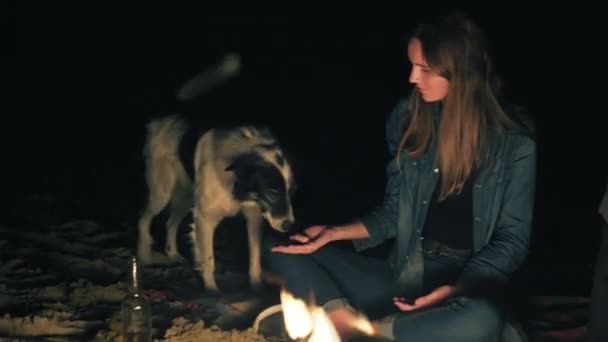 Приваблива біла жінка годує милого собаку, сидячи біля багаття пізно вночі. Крупним планом добра жіноча рука з шматочком ковбаси і обличчям грайливої собаки — стокове відео
