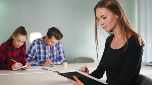 Porträt einer jungen Studentin, die ein Buch liest und an ihren Hausaufgaben arbeitet, während ihre Freunde im Hintergrund studieren. Zeitlupenschuss — Stockvideo