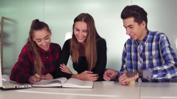 Estudiantes sonrientes trabajando en sus tareas sentados juntos en la mesa y tomando café. Grupo de jóvenes en el encuentro en apartamento moderno. Disparo en cámara lenta — Vídeo de stock