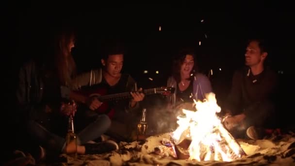 Gruppo multirazziale di giovani ragazzi e ragazze seduti vicino al falò a tarda notte a cantare canzoni e suonare la chitarra. Allegro afroamericano che suona la chitarra per i suoi amici — Video Stock