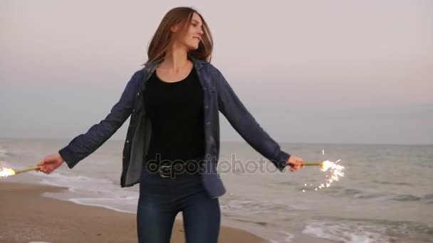 Giovane bella donna che corre in riva al mare durante il tramonto e tiene accese candele scintillanti in entrambe le mani, girandosi, sollevandole. Colpo di rallentamento — Video Stock