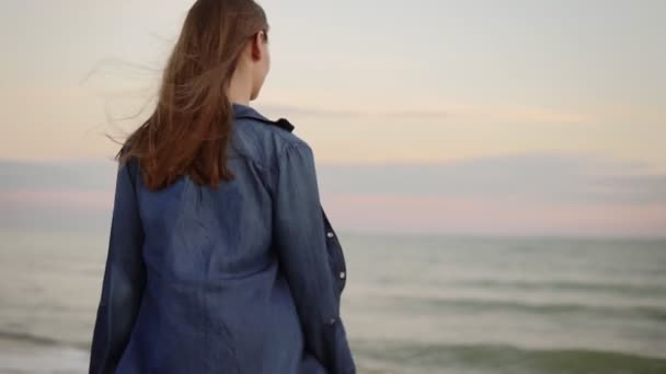 Visão traseira da mulher atraente chegando ao mar e experimentando a temperatura da água com a mão na praia durante o pôr do sol. Tiro em câmara lenta — Vídeo de Stock