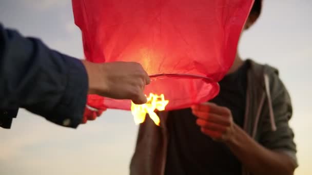 Closeup view of young multiethnic couple holding red paper lantern before launching. Attractive woman with her african boyfriend during romantic date at the beach during sunset — Stock Video