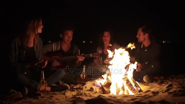 Alegre afroamericano tocando la guitarra para sus amigos junto al fuego. Grupo multirracial de niños y niñas sentados junto a la hoguera a altas horas de la noche cantando canciones y tocando la guitarra — Vídeos de Stock