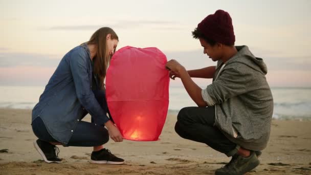 Linda pareja multiétnica sosteniendo linterna de papel rojo antes de su lanzamiento. Mujer atractiva con su novio africano sosteniendo linterna con fuego antes de dejarlo volar. Cita romántica en la playa al atardecer — Vídeo de stock