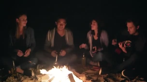 Vista de perto da fogueira à noite. Jovens sentados ao lado do fogo à noite, tocando guitarra. Amigos alegres cantando músicas, conversando e se divertindo juntos — Vídeo de Stock