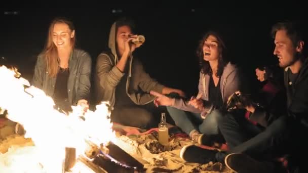 Il falò a tarda notte: giovani allegri seduti accanto al fuoco la sera, che suonano la chitarra e bevono birra. Amici allegri che cantano canzoni, parlano e si divertono insieme — Video Stock