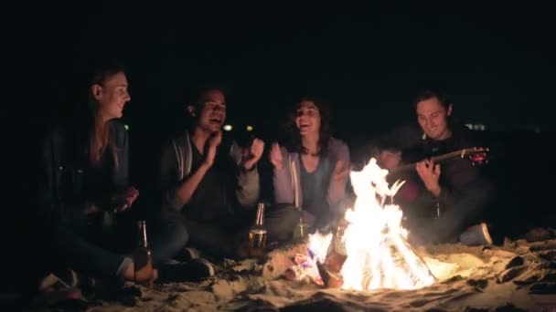 Grupo multiétnico de jovens sentados à beira da fogueira à noite e cantando canções e tocando guitarra, batendo palmas — Vídeo de Stock