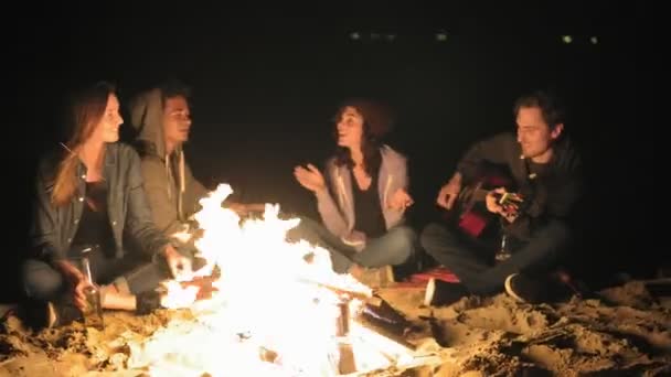 Κάμερα κινείται γύρω από τη φωτιά, αργά τη νύχτα: νέοι χαρούμενα συνεδρίαση από τη φωτιά το βράδυ, παίζει κιθάρα και πίνοντας μπύρα. Χαρούμενα φίλους τραγουδούν τραγούδια, μιλώντας και διασκεδάζοντας — Αρχείο Βίντεο