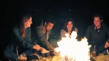 Grup çok ırklı gençlerin Tarih hatmi kavurma ateşte birlikte sahilde gece geç saatlerde, bira içmek sopa