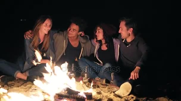 Diverso grupo de personas sentadas juntas junto al fuego tarde en la noche y abrazándose, cocinando salchichas y bebiendo cerveza. Amigos alegres hablando y divirtiéndose juntos — Vídeos de Stock