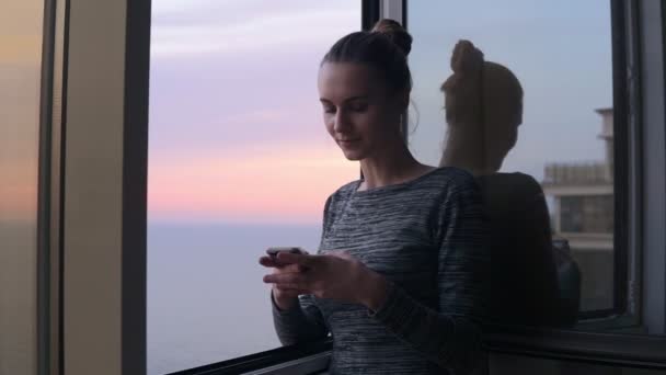 Krásná mladá žena sedí doma u otevřeného okna s úsměvem při pohledu na telefon a píše zprávu během západu slunce. Krásná obloha na pozadí — Stock video
