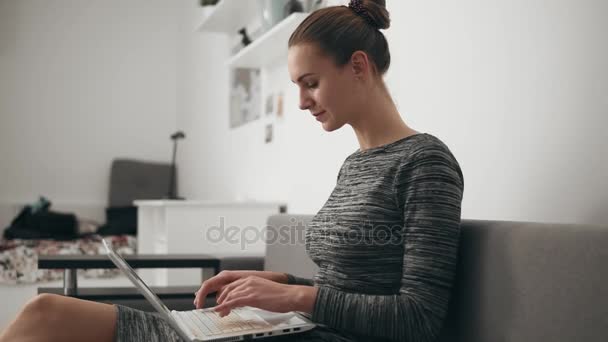 Vista laterale della giovane donna a casa seduta sul divano, che lavora con un computer portatile e digita testo guardando velocemente lo schermo — Video Stock