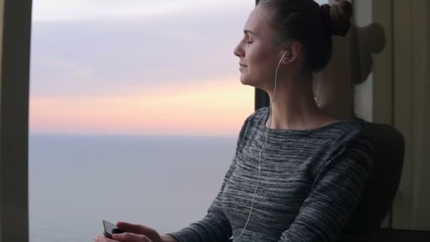 Chica sonriente parada junto a la ventana durante una puesta de sol y escuchando música con auriculares usando el teléfono móvil. Disfrutando de la música en cámara lenta — Vídeo de stock