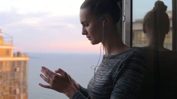 Lachende meisje permanent door het venster tijdens een zonnige dag en luisteren naar muziek met hoofdtelefoons gebruik mobiele telefoon — Stockvideo