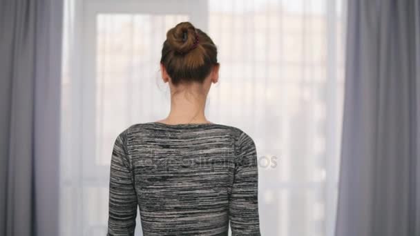 Vista posterior de la joven mujer atractiva abriendo cortinas en una ventana grande y dejando que la luz en la habitación. Mirando por la ventana. Disparo en cámara lenta — Vídeo de stock