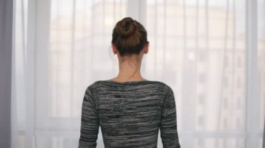 Arkadan Görünüm pencere örtüsünü açmak perdeleri geliyor ve pencereden dışarı bakıyor genç kadın. Manzarayı dışında. Slowmotion atış
