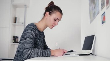 Masaya oturan ve onun kalemi bir mektup yazma güzel ve genç birbayan. Tablo açık laptop. Bir form veya bir belge doldurma notlar bir defterde yapma işkadını