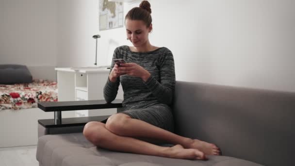 Рух круглої камери: усміхнена молода жінка сидить на дивані вдома, дивлячись на телефон і набираючи повідомлення — стокове відео