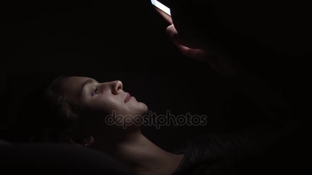 Vista de cerca de una mujer acostada en un sofá y usando su teléfono en una habitación oscura, charlando en la oscuridad con un teléfono inteligente, comunicándose con amigos en línea — Vídeos de Stock