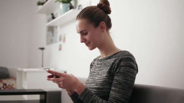 Krásná mladá žena sedí na gauči doma s úsměvem při pohledu na telefon a píše zprávu — Stock video