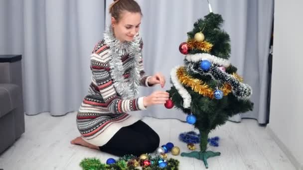 Příprava na Vánoce doma: mladá šťastná žena zdobí vánoční stromeček, závěsné hračky a usmívá se. Snímek v rozlišení 4k