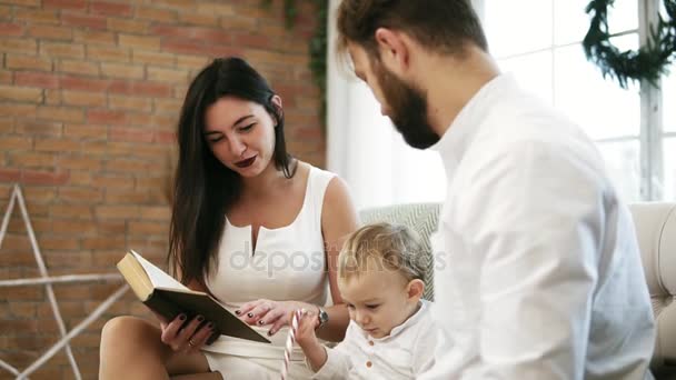Mladá matka čtení kniha pro její syn a manžel. Rodinné čtení Vánoční příběh při posezení u vánočního stromu. Šťastný chlapec zatleskal. Silvestr s milující rodinu — Stock video