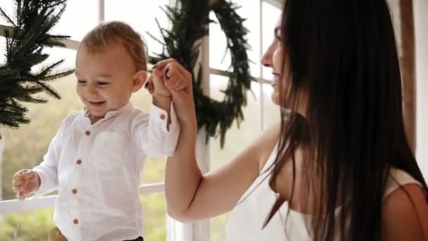 Jonge moeder houdt haar zonen handen terwijl hij op de vensterbank versierd met de kroon van Kerstmis loopt. Gelukkige familie kussen — Stockvideo