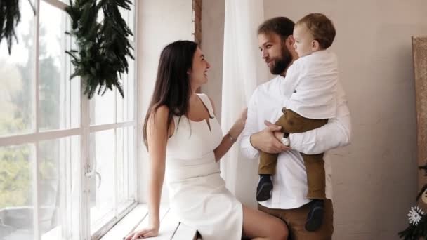 若い父親は、白いドレスの魅力的な母親が窓枠に座っている間窓のそばに立って、彼の男の赤ちゃんを保持しています。ウィンドウにはクリスマス リースが飾られています。愛する家族 — ストック動画