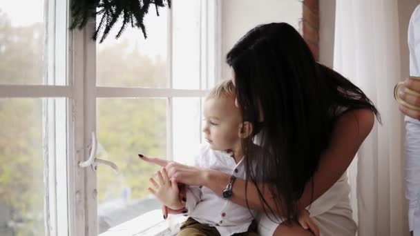 Young mother is sitting with her son on the window sill decorated with Christmas wreath and looking outside. They are talking and smiling. Happy family at home — Stock Video
