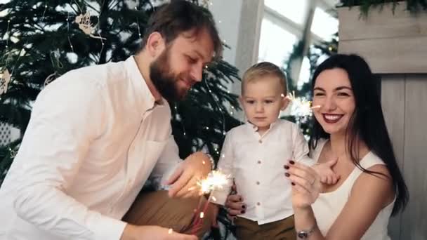 Madre y padre sosteniendo chispeante, mientras que su lindo hijo está mirando la luz. Familia feliz y fuego de Bengala en casa junto al árbol de Navidad. Preciosa familia celebrando la Navidad en casa — Vídeos de Stock