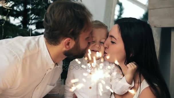 Vackra family jublar det nya året och lampor tomtebloss, unga föräldrar kysser sin härliga barn sitter vid granen — Stockvideo