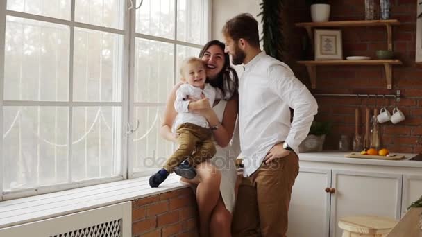 Los padres jóvenes con su lindo hijo sentado en el alféizar de la ventana juntos abrazándose y besándose. La ventana está decorada con guirnalda de Navidad. Amar a la familia juntos en casa — Vídeos de Stock